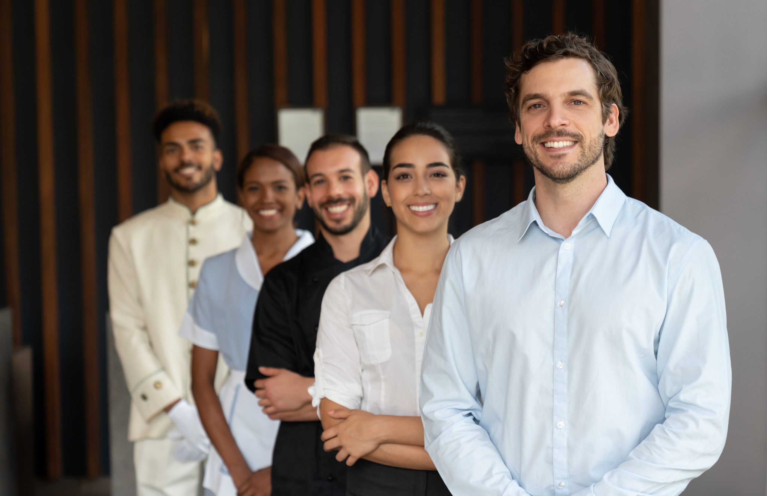 Hotel-manager-and-his-team-standing-behind-all-looking-at-camera-smiling-1068158450_6920x4483